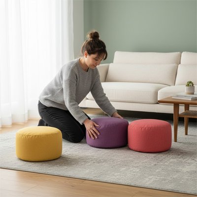 Pouf​ repose pied Femme disposant trois poufs ronds colorés jaune, violet et rouge dans un salon lumineux près d’un canapé.