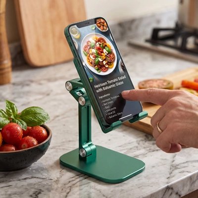 Support pour tablette Main utilisant un smartphone posé sur un support réglable vert, sur un plan de travail de cuisine près de tomates et de basilic
